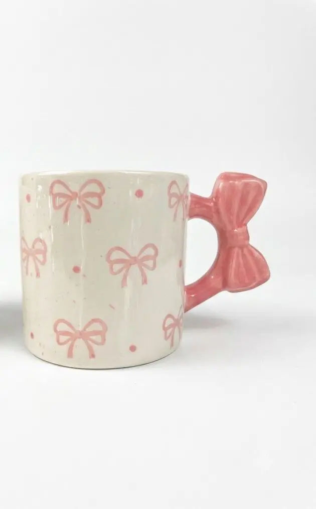 White ceramic mug with a pink bow pattern and a 3D bow-shaped handle.